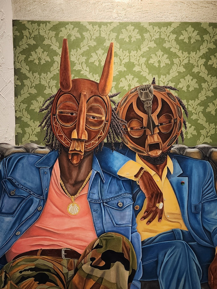 two men of color sitting in relaxed fit clothing sitting on a couch while wearing traditional African masks; behind them is green patterned wallpaper